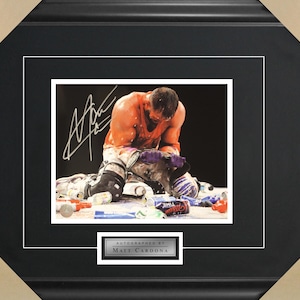 May include: A black and white photo of a wrestler in a red and orange shirt, kneeling on the ground with a white signature above him. The signature reads "Matt Cardona".