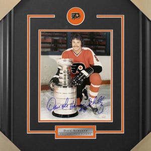 May include: Framed photo of Dave Schultz, a Philadelphia Flyers hockey player, holding the Stanley Cup. The photo is in a black frame with orange and white accents. The player is wearing an orange and black Flyers jersey. The photo includes his signature and a nameplate.