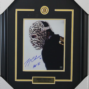 Peut inclure: Une photo encadrée de Gerry Cheevers, un ancien gardien de but de la LNH pour les Bruins de Boston. La photo montre Cheevers portant son masque emblématique avec un design cousu. La photo est signée par Cheevers et comprend le texte "HOF 85".