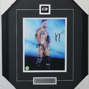 May include: A black framed photo of a wrestler in a wrestling ring. The wrestler is wearing a black and white patterned scarf and black wrestling trunks. The photo is signed by the wrestler, Maxwell Jacob Friedman, with the text "The Salt Of The Earth".