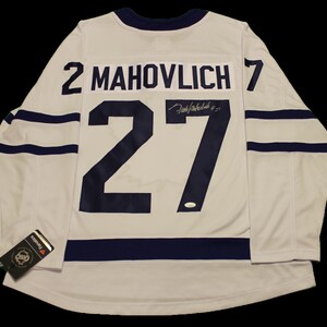 May include: White hockey jersey with blue and white stripes. The number 27 is printed on the back with the name "MAHVOLICH" above it. The jersey is signed by "Darryl Sittler".