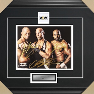 May include: Framed photograph featuring three muscular men, autographed in gold. The image is set against a black background, with the AEW logo at the top. The frame is black with a white matting, and the bottom has a silver plate that reads "AUTOGRAPHED BY SCU."