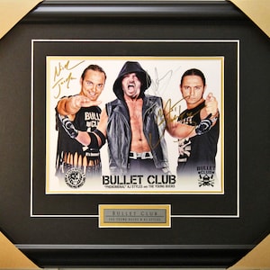 May include: A framed photograph of three wrestlers, signed by each of them. The wrestlers are wearing black clothing and have a serious expression on their faces. The photo is titled "Bullet Club" and features the text "Phenomenal AJ Styles and the Young Bucks".