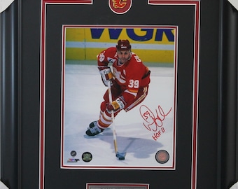 Puede incluir: Fotografía enmarcada de Doug Gilmour con la camiseta de los Calgary Flames, número 39, sobre el hielo. La foto está en un marco negro con un paspartú rojo y blanco. El logotipo de los Calgary Flames está en la parte superior y una placa en la parte inferior dice "Doug Gilmour, Calgary Flames."