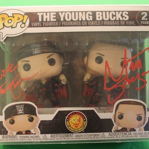 May include: A two-pack set of The Young Bucks wrestling figures in a clear plastic box. The figures are dressed in red and black wrestling attire. The figures are signed by the wrestlers. The box has a warning label about choking hazards.