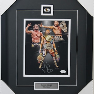 May include: A black framed photo of three wrestlers in a ring. The wrestlers are wearing wrestling gear and holding championship belts. The photo is signed "Kenny Omega "The Elite" ".