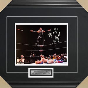 May include: A black and white photo of two wrestlers in a wrestling ring. The wrestler on top is in mid-air, with his leg raised, and the wrestler on the bottom is lying on the mat. The wrestler on top has signed the photo "El Hijo del Vikingo".