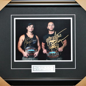 May include: A framed photograph of two men, both wearing black tank tops and holding championship belts. The men are smiling and have signed their names in gold ink on the photo. The text "The Young Bucks" and "Nick & Matt Jackson" is printed below the photo.