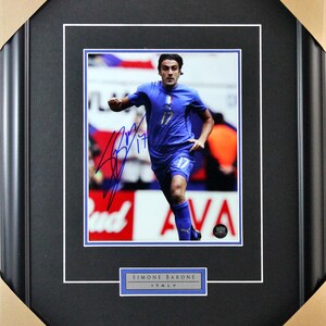 May include: A framed photograph of a soccer player in a blue jersey with the number 17 on it. The player is running with the ball and has signed the photo. The photo is framed in a black frame with a white mat. The player's name, Simone Barone, and the word Italy are printed below the photo.