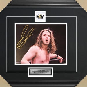 May include: A black and white photo of a wrestler with long hair, wearing a black wrestling strap on his wrist. The photo is signed in gold ink with the name "Joey Janela". The photo is framed in a black frame with a gold "AEW" logo in the top right corner.