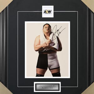 May include: A black and white framed photo of a wrestler in black and white wrestling shorts. The wrestler is holding a white towel and has signed the photo "Samoa Joe". The photo is framed with a black mat and a silver plaque that reads "Autographed by Samoa Joe".
