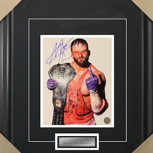 May include: A wrestler in a red and white tank top holds a championship belt with a purple glove on one hand and a finger pointed up. The wrestler has a beard and a serious expression. The image is signed "AUTOGRAPHED BY MATT CARDONA".