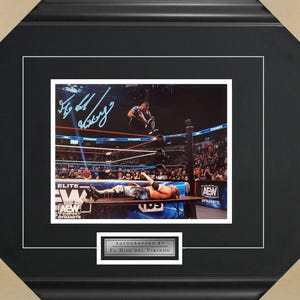 May include: A black and white photo of a wrestler performing a move in a wrestling ring. The wrestler is wearing a blue and black wrestling outfit. The photo is signed by "El Hijo del Vikingo".