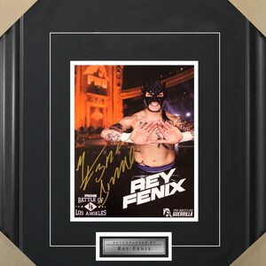 May include: A black and white photo of a wrestler wearing a black mask and a purple and black wrestling outfit. The wrestler is making a hand gesture. The photo is signed by the wrestler, Rey Fenix, and has the text "Rey Fenix" in gold letters. The photo is framed in a black frame with a white mat.