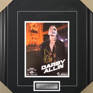 May include: A black and white photo of a man with a skull face paint, wearing a black jacket and a necklace. The photo is framed in a black frame with a white mat. The text "Darby Allin" is written in gold letters on the photo. The text "Pro Wrestling Guerrilla" is written in white letters on the photo. The text "Autographed by Darby Allin" is written in silver letters on a black background.