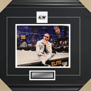 May include: A black and white photo of a wrestler sitting on a wrestling ring apron. The wrestler is wearing a denim jacket and jeans. The photo is framed in a black frame with a gold "AEW" logo in the top right corner and a gold "AUTOGRAPHED BY ORANGE CASSIDY" plaque in the bottom center.
