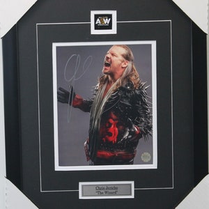 May include: Framed photo of Chris Jericho, "The Wizard", in a black leather jacket with silver spikes. The photo is signed and features the AEW Wrestling logo. The frame is black with a white mat.