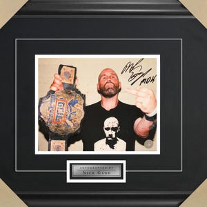 May include: A black and white photo of a man holding a wrestling championship belt. He is wearing a black t-shirt with a skull graphic and is giving the middle finger. The photo is signed "Nick Gage" in black ink.