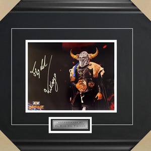 May include: A black and white photo of a wrestler wearing a horned helmet and a black and silver costume. The wrestler is holding a championship belt. The photo is signed "El Hijo del Vikingo" in white ink. The photo is framed in a black frame with a silver mat.