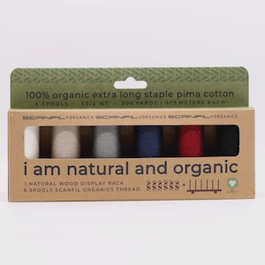May include: A rectangular cardboard box containing six spools of Scanfil Organics thread in white, beige, gray, navy, red, and black. The packaging text reads 100% organic extra long staple pima cotton, 500 yards, and i am natural and organic.