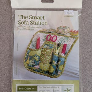 May include: A sewing pattern package titled The Smart Sofa Station, a multi-craft sofa organiser. The cover displays a quilted fabric organiser with pockets holding scissors, pens, and tools. It includes a pin cushion and lists a finished size of 22 inches by 9.5 inches.