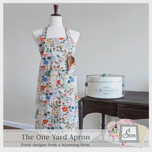 May include: A floral print apron on a mannequin with a wooden spoon in the pocket. The apron has a white background with red, blue, and yellow flowers. A white cake carrier with the word "PATISSERIE" sits on a dark wooden table. The text reads "The One Yard Apron."