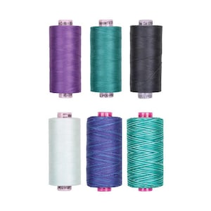 May include: Six cylindrical spools of sewing thread arranged in two rows of three. The top row features solid purple, teal, and dark gray thread. The bottom row includes solid white, variegated blue and purple, and variegated teal and white thread.