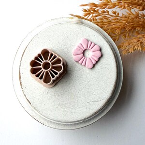 May include: Two pink flower-shaped cookie cutters and a flower-shaped fondant. The cutters are square with a detailed floral design. The fondant is a smaller, solid pink flower shape. The items are on a white, distressed, round surface.