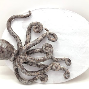 May include: A ceramic octopus sculpture in shades of gray and brown, mounted on a white, textured, circular base. The octopus has detailed tentacles with curled ends, creating a three-dimensional effect. The sculpture is a decorative art piece.