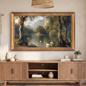 May include: A rectangular landscape painting in a gold frame depicting a lake with white swans, trees, and mountains, mounted above a long, slatted wooden media console with two cabinets and an open centre shelf containing books and a white vase.
