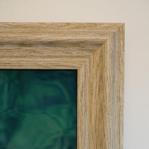 Pode incluir: Uma obra de arte emoldurada com uma moldura de madeira clara. A obra de arte dentro da moldura apresenta um design abstrato verde e azul. A moldura é retangular e a obra é exibida em uma parede branca. As dimensões da moldura são em centímetros.