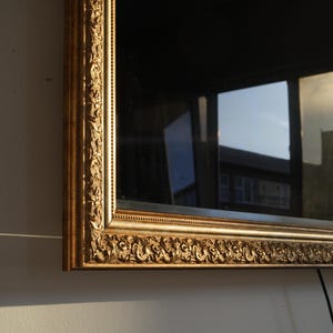May include: A rectangular mirror with an ornate, gold-coloured frame. The frame features detailed scrollwork and a decorative border. The mirror reflects a view of buildings and a cloudy sky, with a black cable visible.