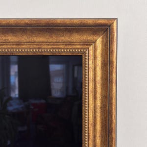 Pode incluir: Um espelho retangular com uma moldura larga e ornamentada com um acabamento dourado antigo desgastado. A moldura apresenta uma superfície texturizada e manchada com tons castanhos escuros e um detalhe de borda perolada no interior.