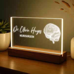 以下が含まれることがあります： 「Dr. Olivia Hayes, Neurosurgeon」の文字と3D脳グラフィックが入った、照明付きのデスクサイン。透明アクリル製で、木製の台座に置かれています。柔らかく温かい光で照らされています。