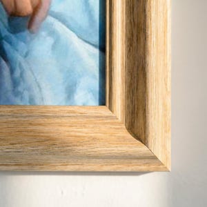 Puede incluir: Primer plano de un marco de fotos de madera marrón claro con un acabado de veta de madera natural. El marco rodea una parte de una pintura con tonos azules y blancos. Se ve la esquina del marco.