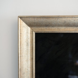 May include: Close-up of a rectangular framed mirror. The frame has a distressed, metallic gold finish. The mirror's surface is black, reflecting a dark space. The frame's corner is visible against a white wall.
