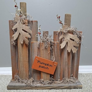 May include: A decorative wooden sign featuring three vertical planks of varying heights, accented with burlap leaf cutouts, dried moss, and small red berries. An orange rectangular tag hangs from the center, displaying the text Pumpkin patch.