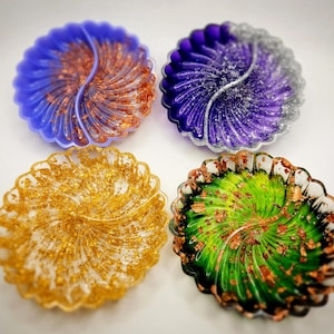 May include: Four decorative resin coasters in a circular shape with a scalloped edge. The coasters are in shades of blue, purple, gold, and green, each with a copper-colored swirl design. The coasters are on a white surface.