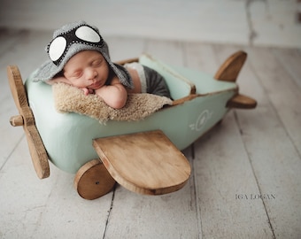 Puede incluir: Un recién nacido dormido con gorro de aviador gris y pantalones cortos, en un avión de juguete de madera azul claro. El avión tiene alas y una hélice de madera. El bebé descansa sobre un material similar a la piel de oveja.