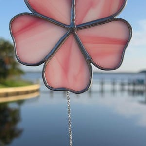 以下が含まれることがあります： ピンクと白の花びらが銀色で縁取られたステンドグラスの花のサンキャッチャー。透明なクリスタルのしずくが、花の下のチェーンから吊り下がっています。背景は、水と空のぼやけた景色です。