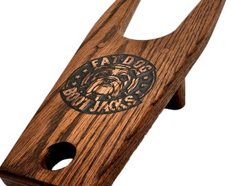May include: A wooden boot jack with a dark brown finish. The boot jack has a slot for the heel and a V-shaped opening to hold the boot. The text "FAT DOG BOOT JACKS" is etched into the wood.