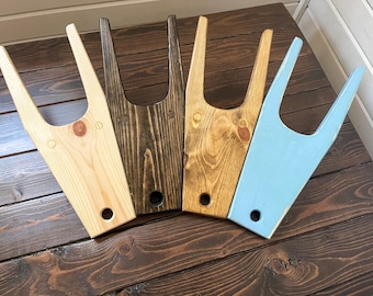 May include: Four wooden boot jacks in different colors: natural wood, dark brown, light brown, and light blue. The boot jacks are all the same shape and size, with a U-shaped opening at the top and a handle at the bottom.
