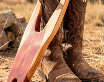 May include: A wooden boot jack with a reddish-brown finish leans against a pair of brown leather cowboy boots. The boots have decorative stitching and are worn. The boot jack is designed to help remove boots.