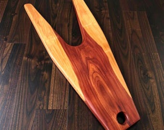 May include: A handcrafted wooden boot jack with a V-shaped design. The boot jack is made of two tones of wood, with a reddish-brown center and lighter-colored edges. It has a hole at the end for hanging. The boot jack is designed to help remove boots.