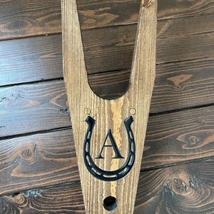 May include: Wooden boot jack with a horseshoe design and the letter "A" engraved on it. The boot jack is stained brown and has a hole in the bottom.