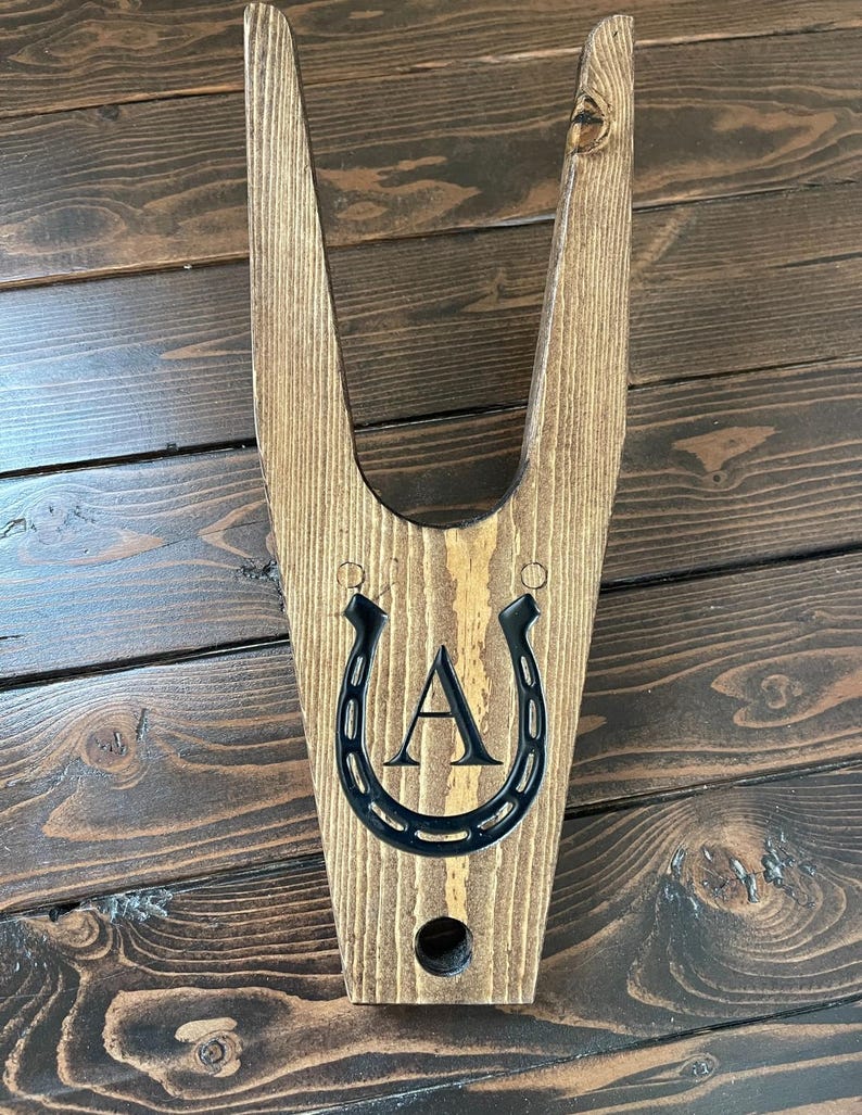 May include: Wooden boot jack with a horseshoe design and the letter "A" engraved on it. The boot jack is stained brown and has a hole in the bottom.