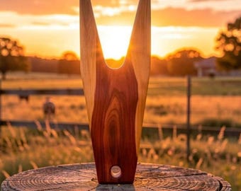 May include: A handcrafted wooden boot jack with a V-shaped design. The boot jack is made of light and dark brown wood, with a hole at the base. The background features a sunset over a field.