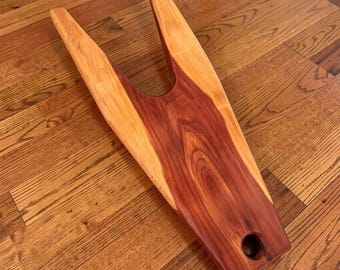 May include: A wooden boot jack with a V-shaped design. The boot jack is made of two tones of wood, with a reddish-brown center and lighter brown edges. A hole is drilled at the end of the jack.