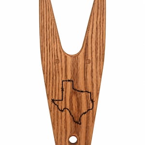 May include: A wooden boot jack with a natural brown wood grain finish. The surface features a laser-engraved outline of the state of Texas, depicted with a barbed wire pattern along the border. There is a circular hole at the bottom center.
