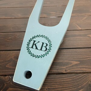 May include: A light blue wooden boot jack with a green wreath and the initials "KB" in the center.
